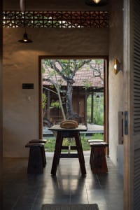 A wooden table with a woven basket sits between wooden stools, in front of a concrete wall with a tiled floor.
