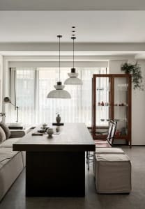 A modern minimalist living room and dining area with a wooden table and chairs, white pendant lights, and a glass-front displ