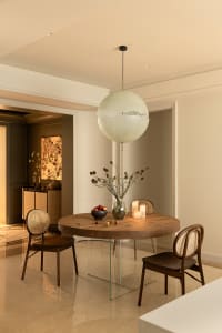 A modern dining room with a round wooden table, glass legs, and a large spherical pendant light hanging above. The space is d