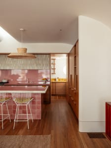 A modern kitchen with wooden floors, pink tiles, and white bar stools.