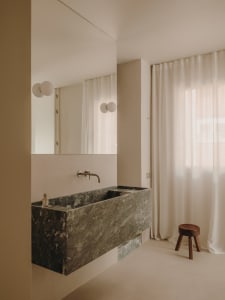 Minimalist bathroom interior with a large, dark marble sink, wall-mounted faucet, and floor-to-ceiling white curtains.