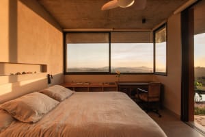 A modern minimalist bedroom with concrete walls, large windows, and a view of the mountains at sunset.