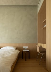 A minimalist bedroom featuring a bed with white bedding, a wooden bedside table, and a chair with a wooden backrest.