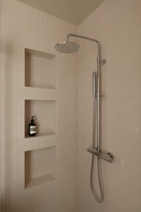 A modern minimalist shower with textured beige tiles, featuring a fixed showerhead and a handheld showerhead mounted on a chr