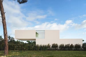 A minimalist, modern residential home with a concrete facade, surrounded by greenery and clear blue sky.