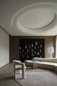 A modern living room with neutral-toned decor, featuring a white sculptural chair, a curved sofa, and a bookshelf filled with