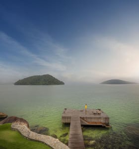 A wooden jetty extending into the ocean with a person standing on it, surrounded by mountains.