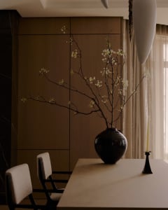 A minimalist living room featuring a wooden table with a vase of blooming branches, two white cushioned chairs, and a candle 