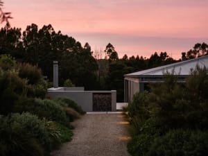 Modern minimalist residential exterior with concrete walls and roof, surrounded by lush greenery at sunset.