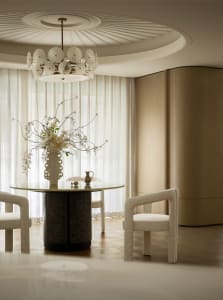 A modern residential dining room featuring a glass table with a vase of flowers, upholstered chairs, and a chandelier.
