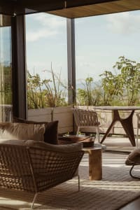 A minimalist living room featuring wooden cushions and rattan furniture, with a view of greenery outside through large glass 