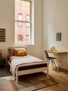 A minimalist bedroom featuring a white bed, colorful pillows, and a wooden desk against a white brick wall.