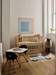 Minimalist children's bedroom featuring a crib, small table, chairs, and a woolly sheep against a window with a plant.
