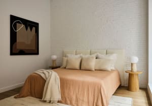 A minimalist bedroom featuring a bed with beige bedding and a wooden headboard, with a modern abstract painting on the wall a