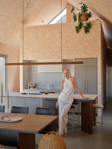 A photograph of a minimalist residential kitchen with wooden accents, featuring a woman leaning on the kitchen island.