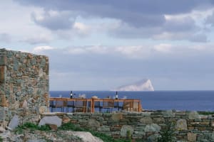 A stone outdoor dining area with a table and chairs overlooking the ocean and a distant island.