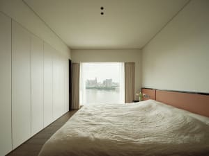 A minimalistic bedroom featuring fabric bedding and wooden accents with a view of the cityscape through the window.