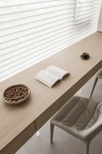 A modern minimalist home office features a wooden desk with an open book, bowl of peanuts, and a mug, alongside a chair and a