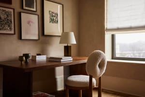 A modern home office setup featuring a wooden desk with a fabric-covered, minimalist chair, framed artworks on the wall, and 
