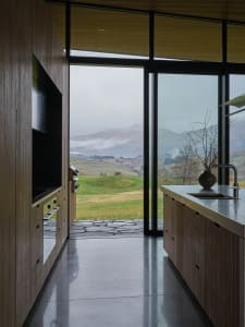 Modern minimalist residential kitchen with large glass windows overlooking a scenic landscape of green hills and mountains.