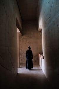 A woman standing in a dimly lit corridor with concrete walls and a wooden beam. She is wearing a black outfit.