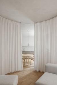 A spacious residence with white curtains, wooden flooring and furniture, and a view into the kitchen area.