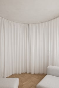 A minimalist living room with white curtains and wooden flooring.