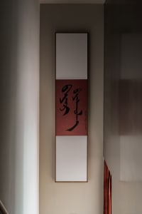 A modern-style calligraphy scroll with a red background and black text hanging on a light-colored wall.