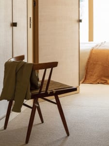 Minimalist room featuring an empty wooden chair in front of a door and a bed with an orange blanket visible in the background