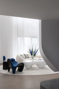 A sleek, modern living room featuring white curtains, a white sofa, and a unique white coffee table with a vase of blue flowe