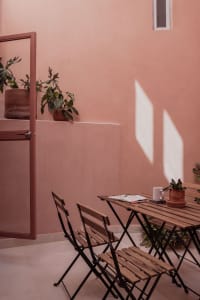 A minimalist patio area with wooden furniture and plants, featuring a pink wall and natural light.