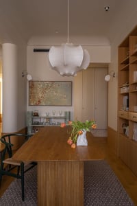 Modern minimalist dining room with wooden table, white pendant light, abstract painting, and bookshelf.