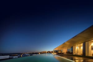 A modern residential home at night, featuring a glass and concrete exterior with an infinity pool.
