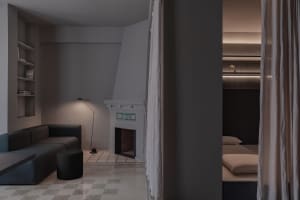 A minimalist bedroom featuring a plaster floor, a bed with beige bedding, and curtains leading to a separate sleeping area wi