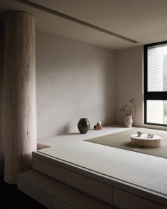 A minimalist living room featuring wooden furniture, a tatami mat, and a small tea set on a wooden platform.