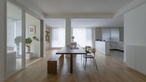 A modern, minimalist residential interior featuring a sleek dining table, chairs, and a kitchen area with white cabinets and 