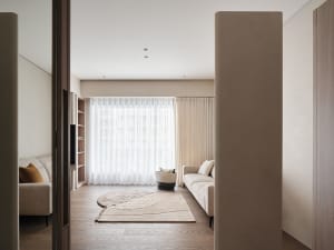 A modern living room featuring neutral-colored furniture, a patterned rug, and large windows with sheer curtains.