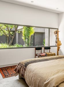 A modern bedroom with a large window overlooking greenery, concrete furniture, and a rug with a traditional pattern on the co