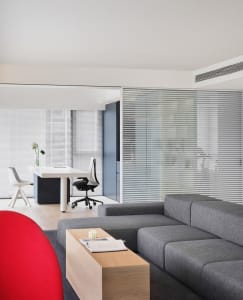 A modern living room featuring a grey sectional sofa, a red armchair, a wooden coffee table, and a white desk with a black ch