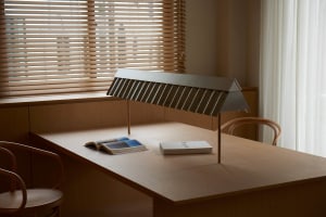 A minimalist, metal desk lamp is placed on a wooden table in a room with wooden blinds and a white curtain.