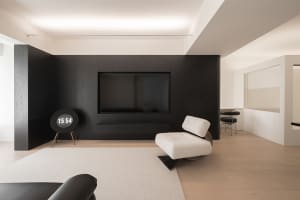 The minimalist living room features black and white decor, with a sleek black wall-mounted TV, white armchair, and wooden flo