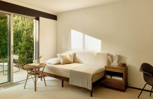 A minimalist bedroom featuring a cream-colored daybed with white and cream bedding, a small wooden side table with books and 