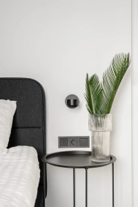 Modern minimalist bedroom featuring a black metal bedside table with a glass vase containing palm leaves.