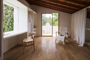 A minimalist room with wooden floors, a white chair, and a glass door leading to a balcony with greenery outside.