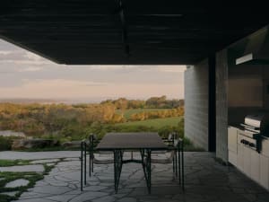 A minimalist outdoor dining area featuring concrete and stone materials, with a table and chairs overlooking a scenic landsca