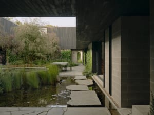 A modern house with a concrete exterior features a garden with water features, pathways, and greenery.