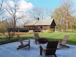 Outdoor deck with wooden chairs and a fire pit surrounded by greenery and a wooden barn in the background.