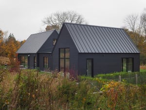 A modern residential building with black metal roofing, concrete siding, and surrounded by greenery and autumn trees.