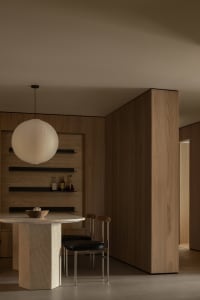 A modern dining room featuring wooden walls and chairs, with a round table, pendant light, and shelves.