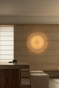 A minimalist interior with a wooden wall featuring a glowing circular light fixture and a wooden dining table with stools.